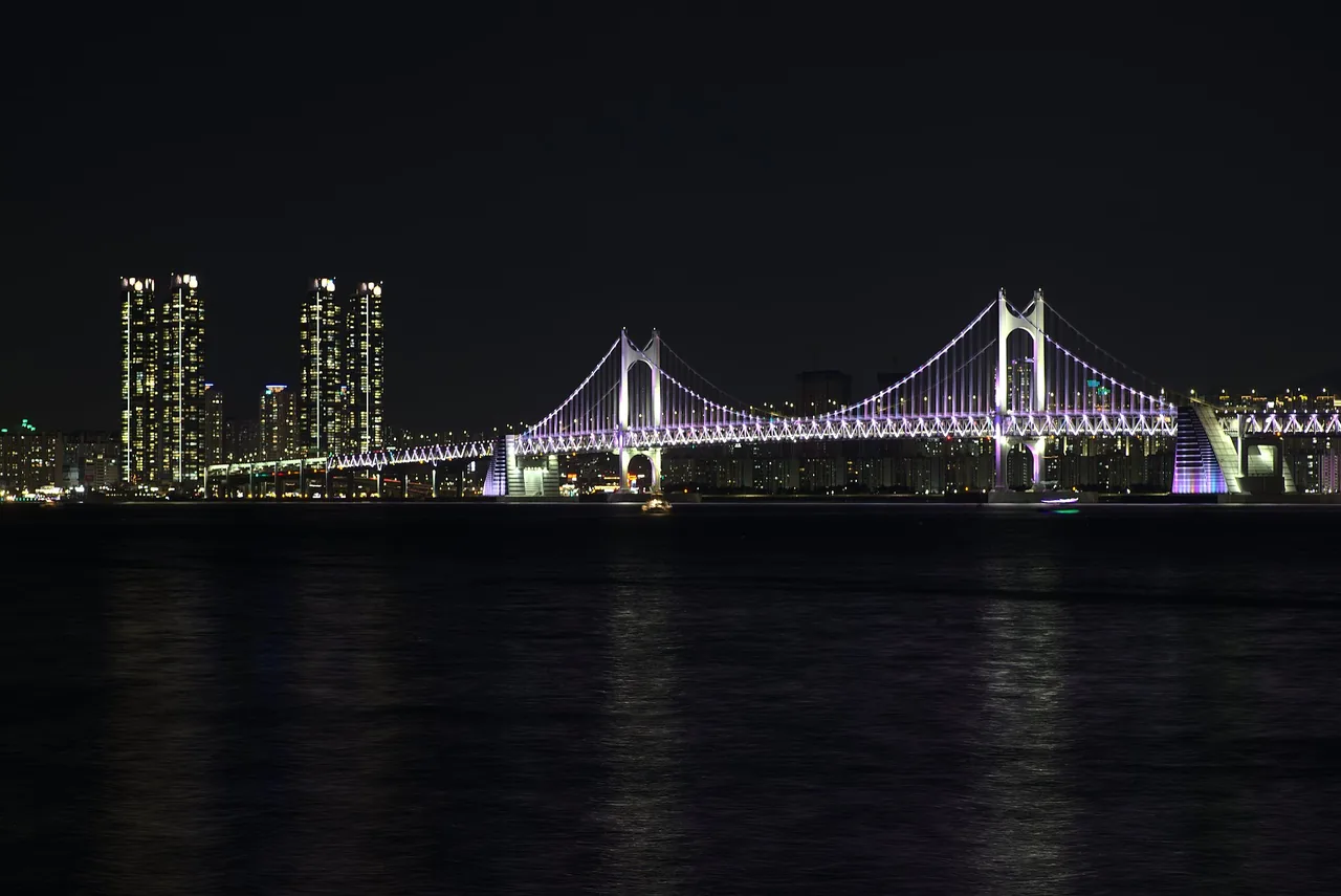 Gwangan Bridge illuminated at night above Gwangalli Beach with the city skyline reflected in the water Gwangan Bridge illuminated at night above Gwangalli Beach with the city skyline reflected in the water