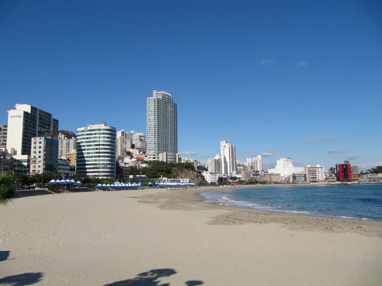 The sandy curve of Songdo Beach with blue parasols and the city skyline, Busan