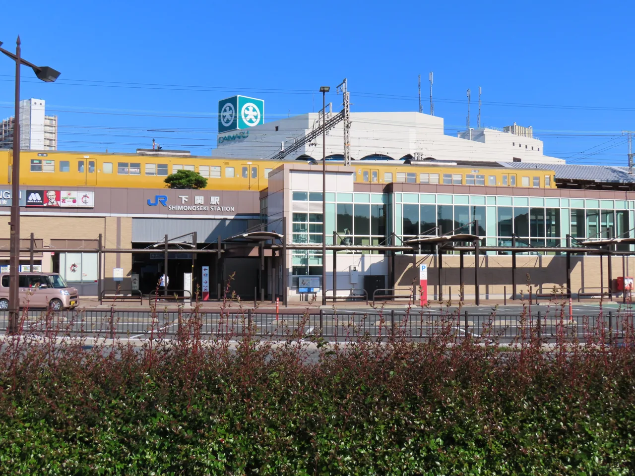 The exterior of JR Shimonoseki Station, with the road in front and a yellow local train standing alongside