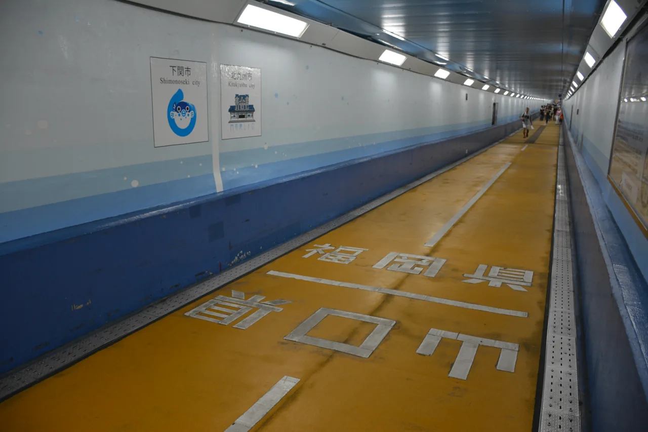 Inside the Kanmon Tunnel pedestrian way, a painted prefectural boundary marking across the floor and walkers further along the tube