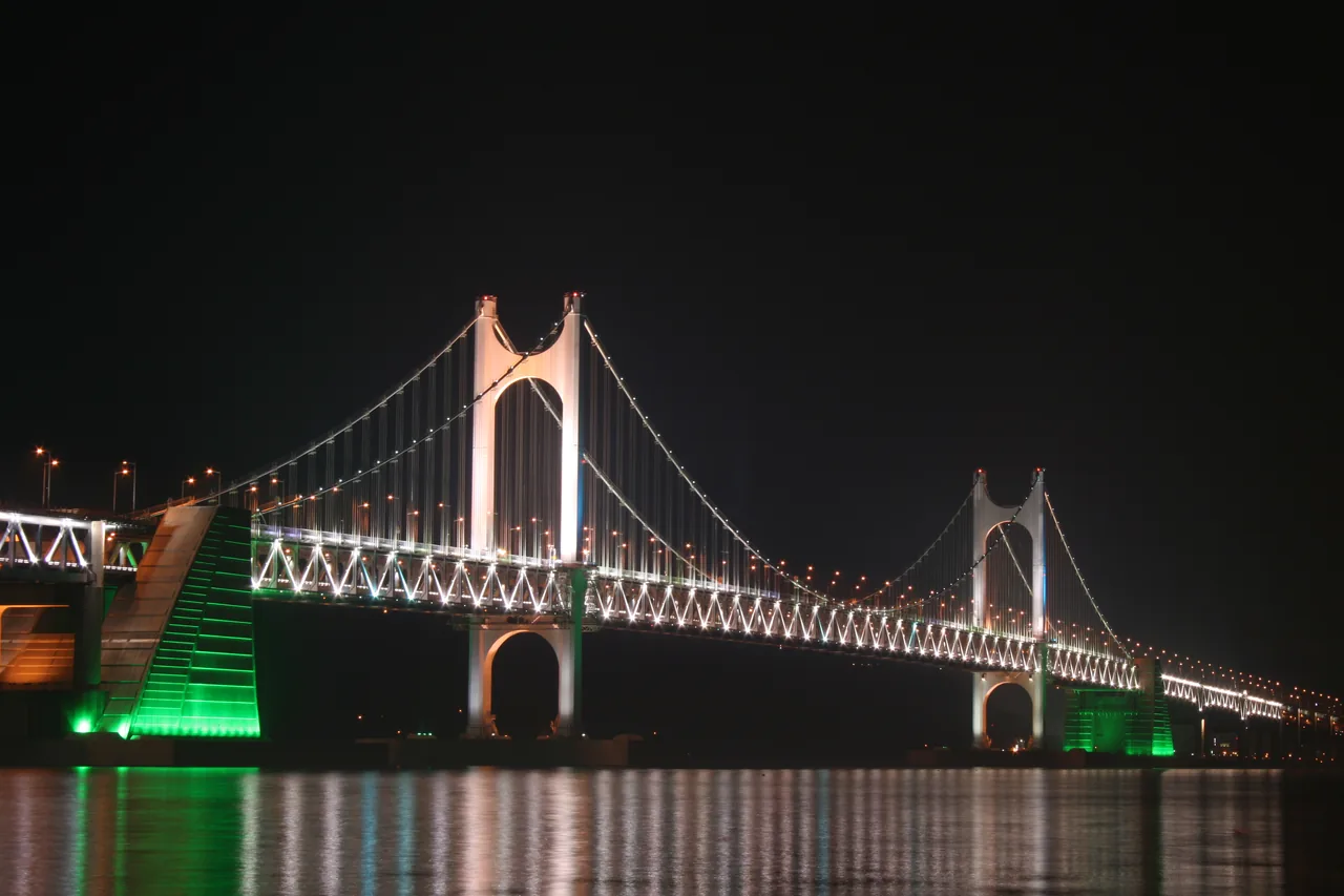 Gwangan Bridge lit at night, illuminated towers and cables above dark water, green light at the pier bases