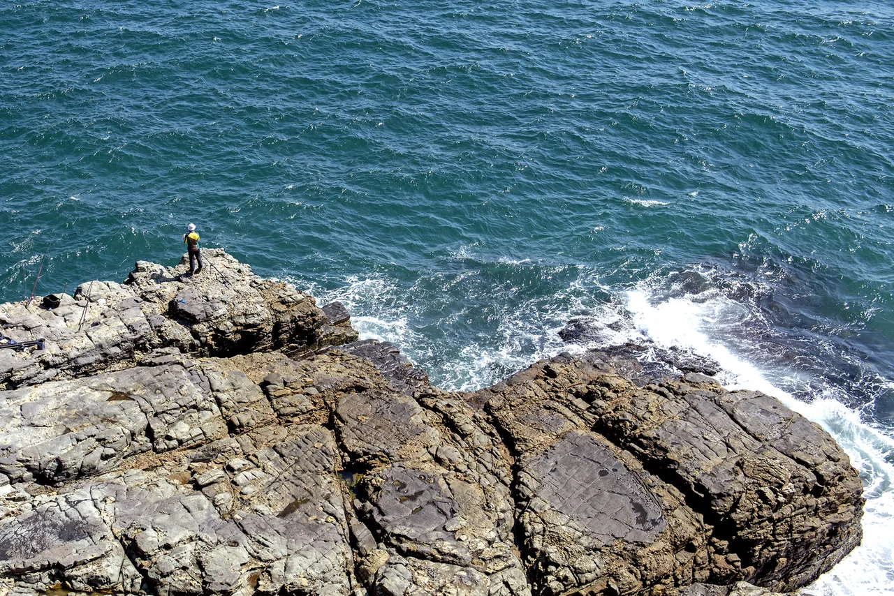A lone angler standing on layered coastal rock as waves break against it