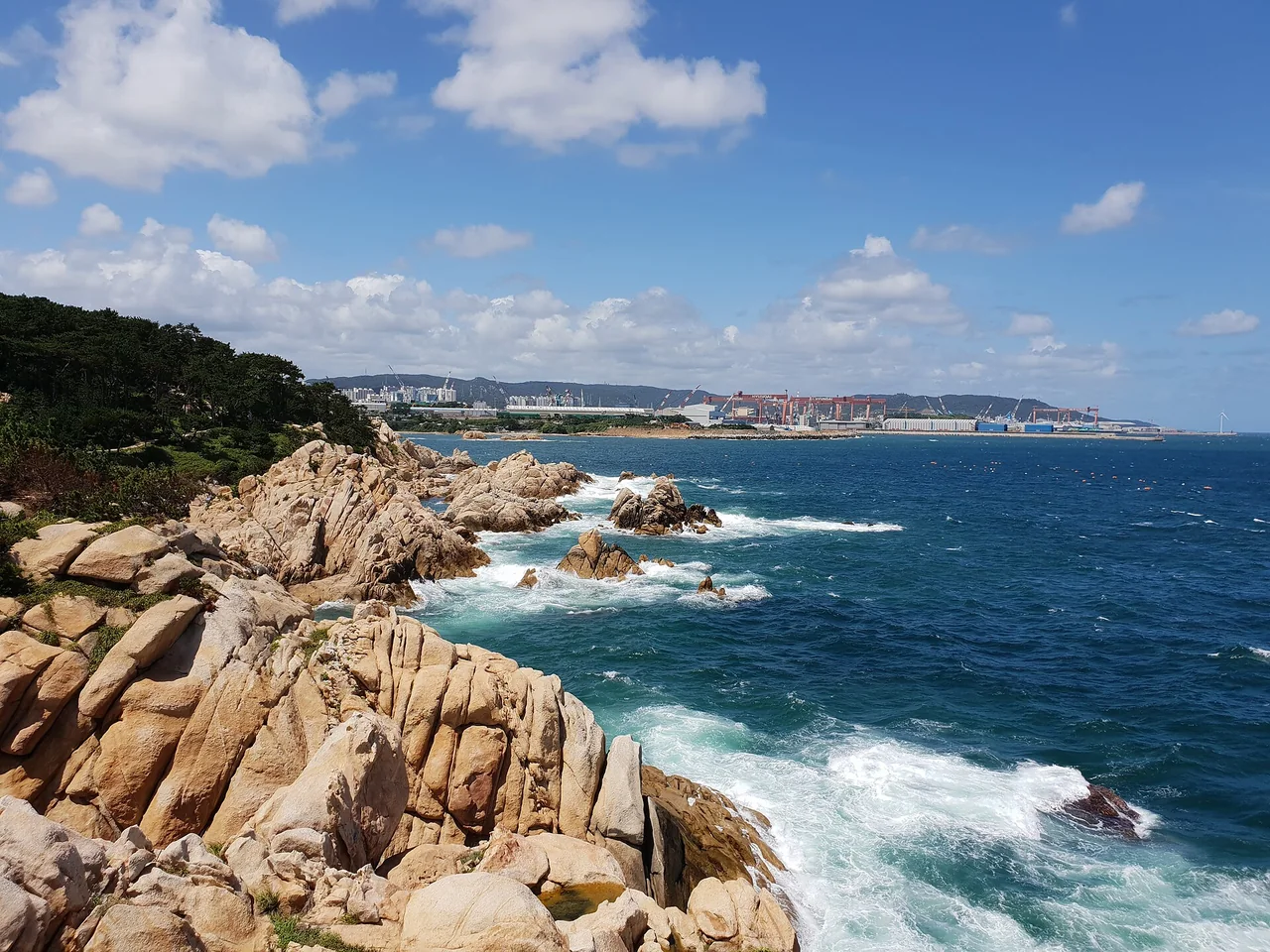 Granite and surf on the Daewangam Park shoreline with shipyards along the horizon opposite