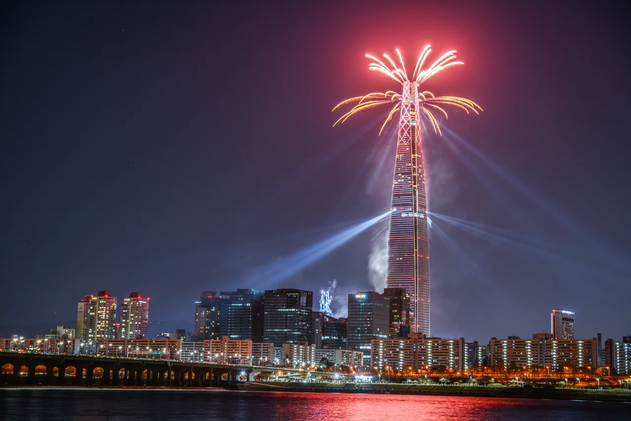 Fuegos artificiales y láseres en lo alto de la Lotte World Tower vista desde el otro lado del río Han, con las luces reflejadas en el agua