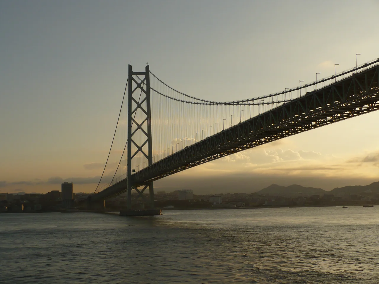 Akashi-Kaikyo Bridge seen from a ship below, its towers and deck in dark silhouette against a golden sky, with a city in the distance