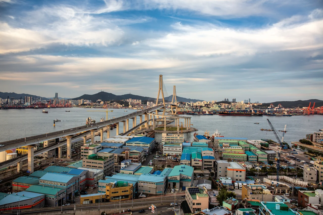 Busan Harbor Bridge, a two-tower cable-stayed bridge, over the sea at Busan's North Port, seen from above with factory and warehouse roofs below and container cranes and mountains in the distance under a cloudy blue sky