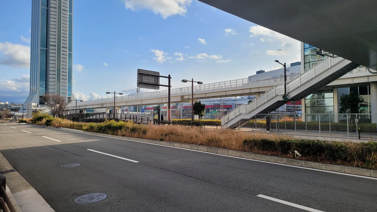 Street near Cosmosquare Station with a tall glass tower on the left, a pedestrian bridge with stairs, an empty road and a blue sky