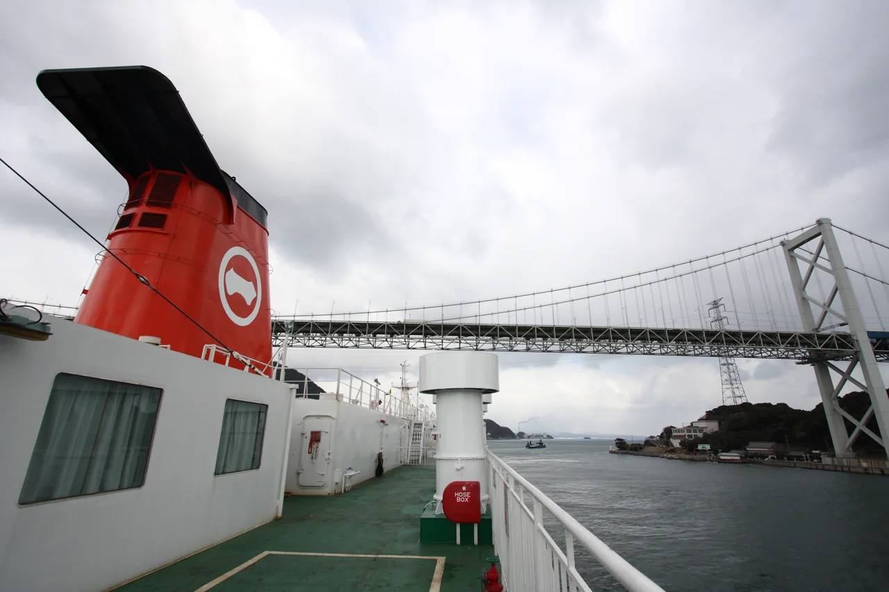 Kanmon Bridge seen from the green deck of a ferry with a red funnel, under a cloudy sky