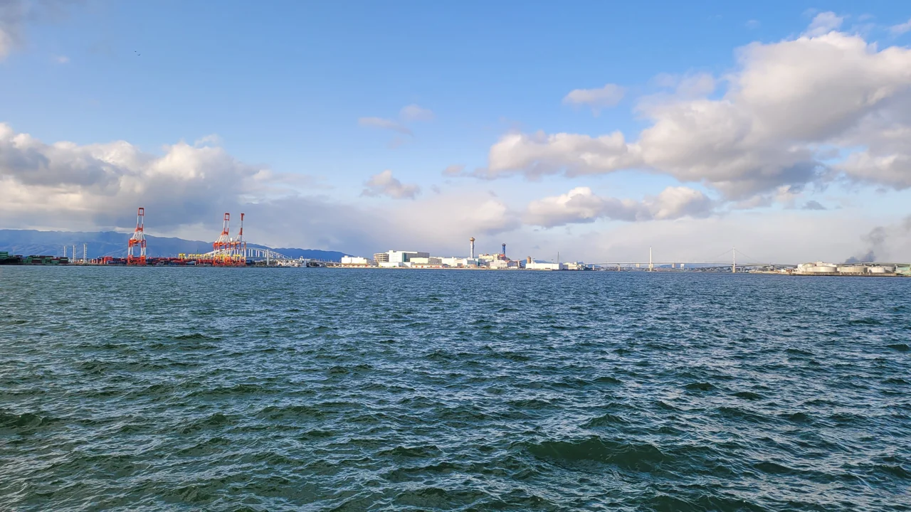 Osaka Port seen across the water from the Cosmosquare side, with red container cranes, white buildings, white clouds and rippling water