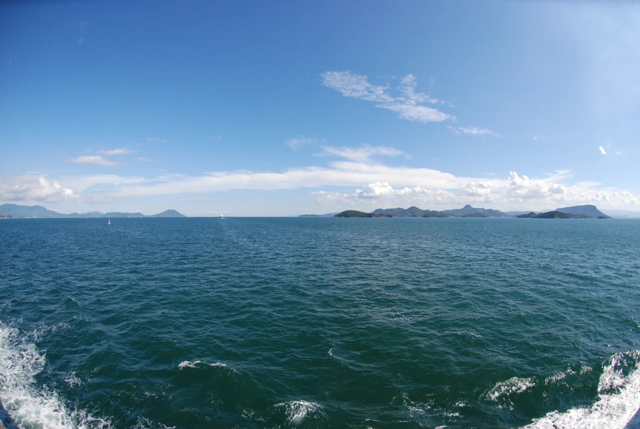 Calm blue sea seen from the stern of a boat, with white wake, distant islands and a blue sky