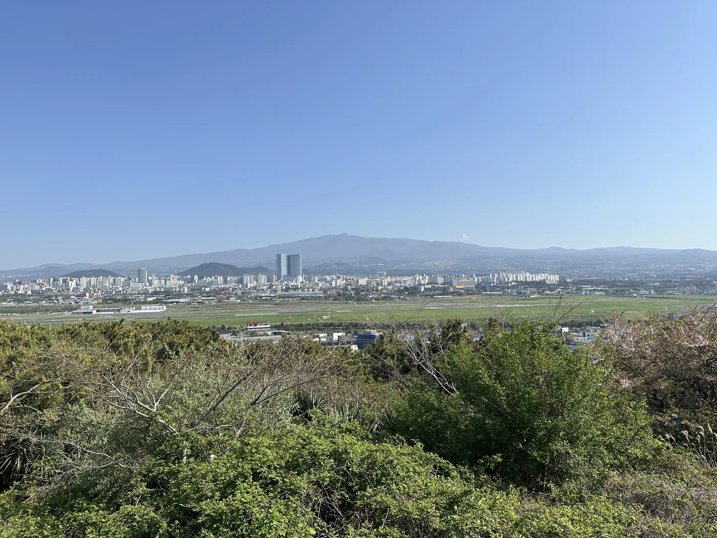 從道頭峰上看機場方向,前景是草叢,橫向展開的跑道與草地,後方是濟州市區建築與高樓,最遠處是漢拏山,天空晴朗 從道頭峰上看機場方向,前景是草叢,橫向展開的跑道與草地,後方是濟州市區建築與高樓,最遠處是漢拏山,天空晴朗