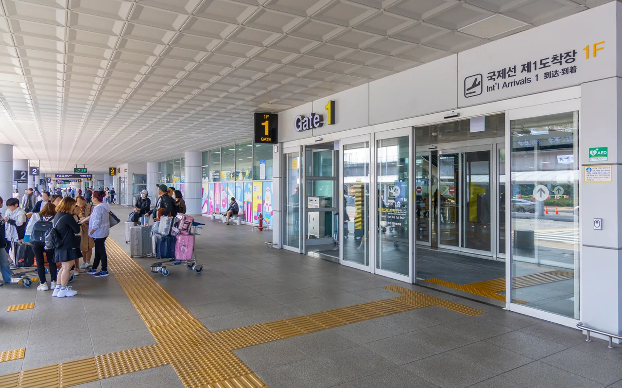 Pavement outside a door of the Gimhae International Terminal marked International Arrivals 1F and Gate 1, with travellers pulling suitcases and yellow tactile paving Pavement outside a door of the Gimhae International Terminal marked International Arrivals 1F and Gate 1, with travellers pulling suitcases and yellow tactile paving