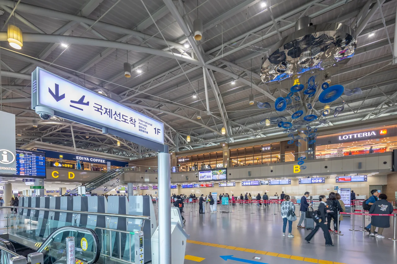 Departures hall of the Gimhae International Terminal with a steel roof, a blue hanging sculpture, a sign for international arrivals on 1F, check-in zone signs B, C and D, and Lotteria and Ediya Coffee signs on the floor above Departures hall of the Gimhae International Terminal with a steel roof, a blue hanging sculpture, a sign for international arrivals on 1F, check-in zone signs B, C and D, and Lotteria and Ediya Coffee signs on the floor above