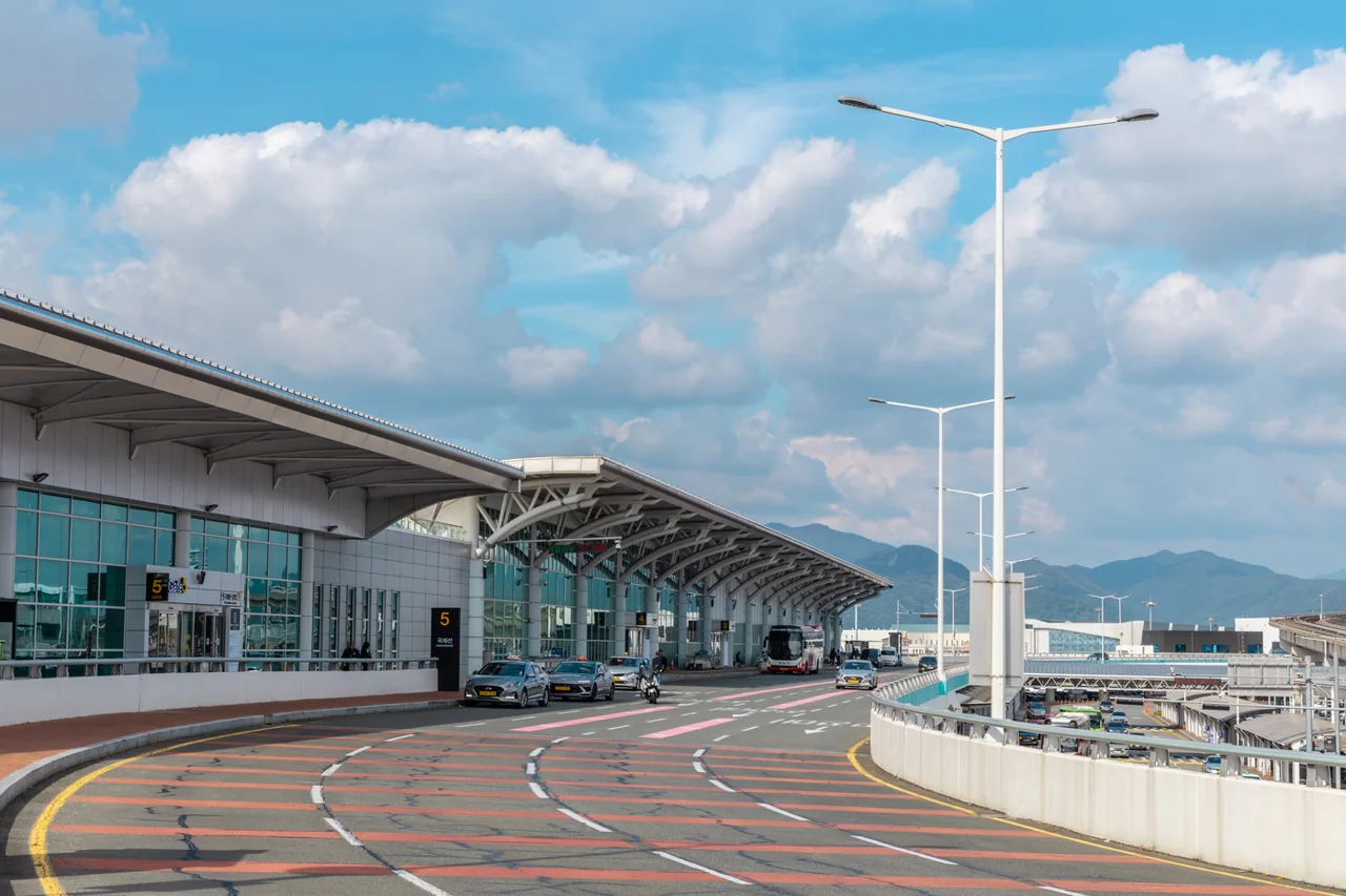 Departures road on the second level of the Gimhae International Terminal, with the glass terminal and curved roof on the left, a gate 5 sign, cars and a bus, and hills under a blue sky Departures road on the second level of the Gimhae International Terminal, with the glass terminal and curved roof on the left, a gate 5 sign, cars and a bus, and hills under a blue sky