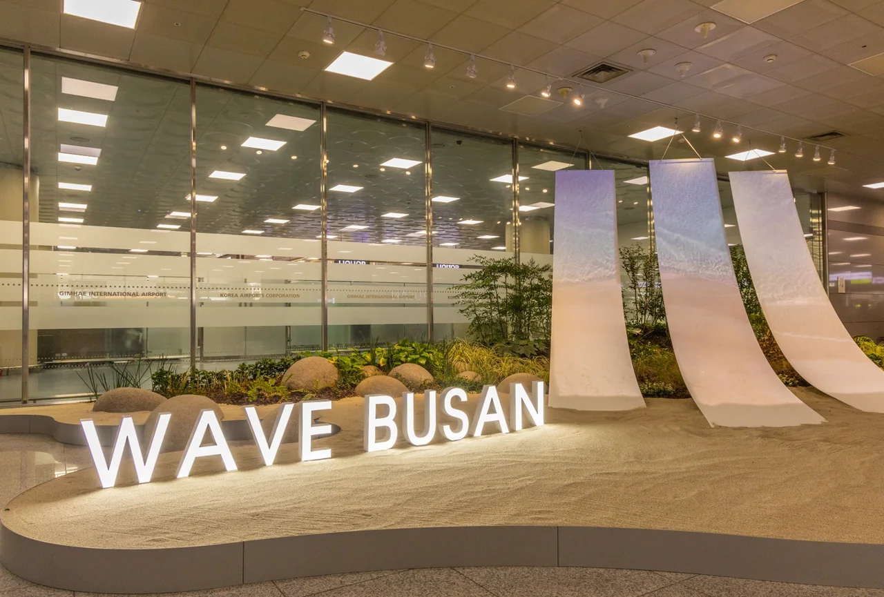 Lit-up WAVE BUSAN letters on an indoor sand garden with three white fabric-like panels and a glass wall behind