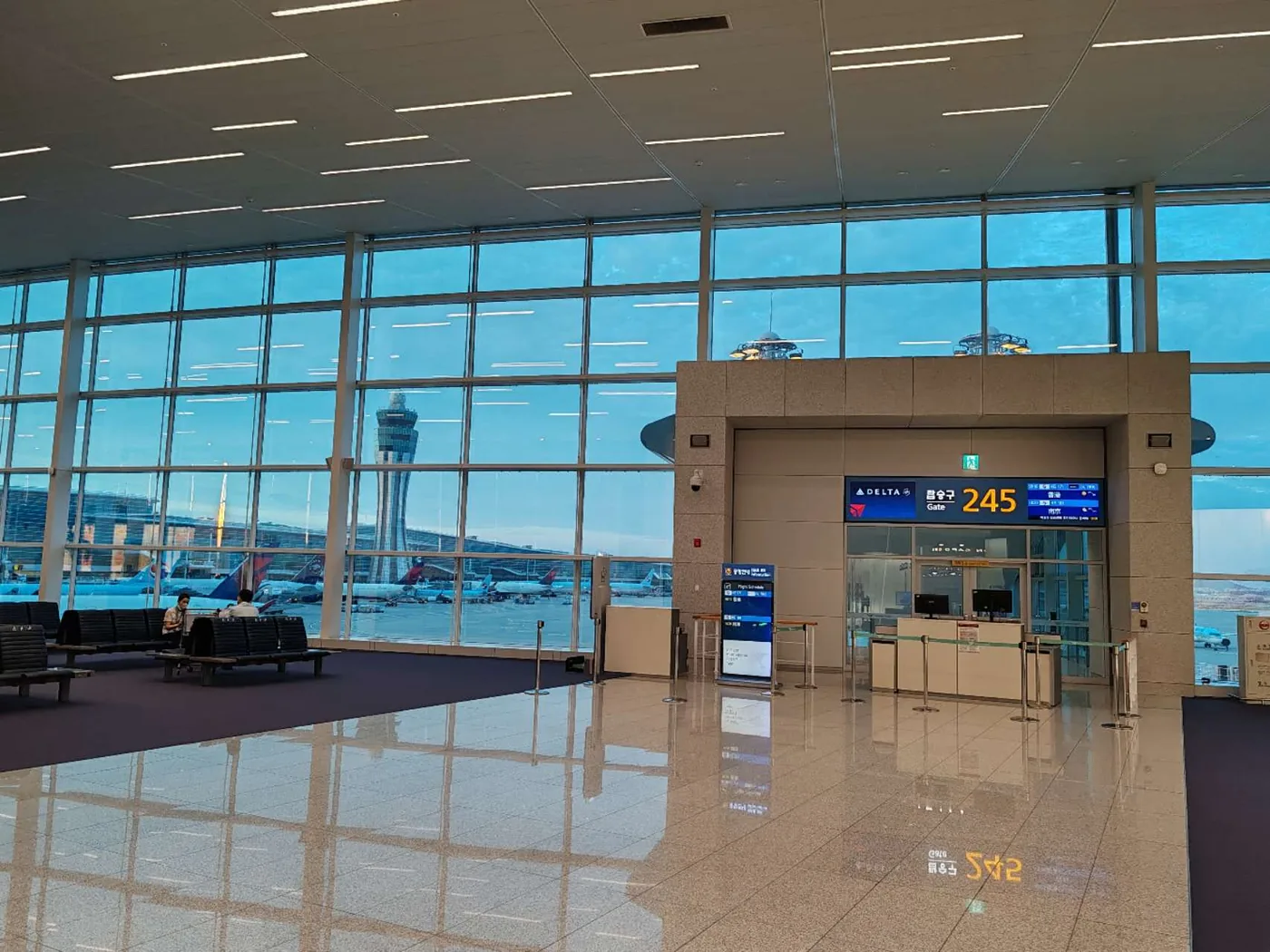 第2旅客ターミナルの245番搭乗口の待合スペース。搭乗カウンターの上に航空会社の表示と「탑승구 245」、空いた椅子の列、ガラス越しに管制塔と駐機場の飛行機、光る床