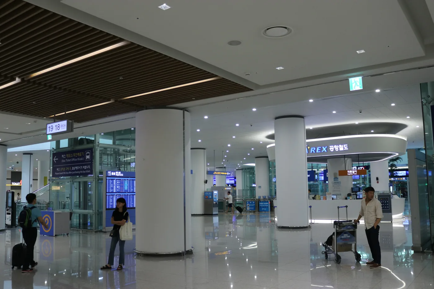 仁川空港2ターミナル駅の待合スペース。丸い「AREX 공항철도」の案内と切符売り場、白い円柱、上の発車時刻の表示、カートを押す人と旅行者