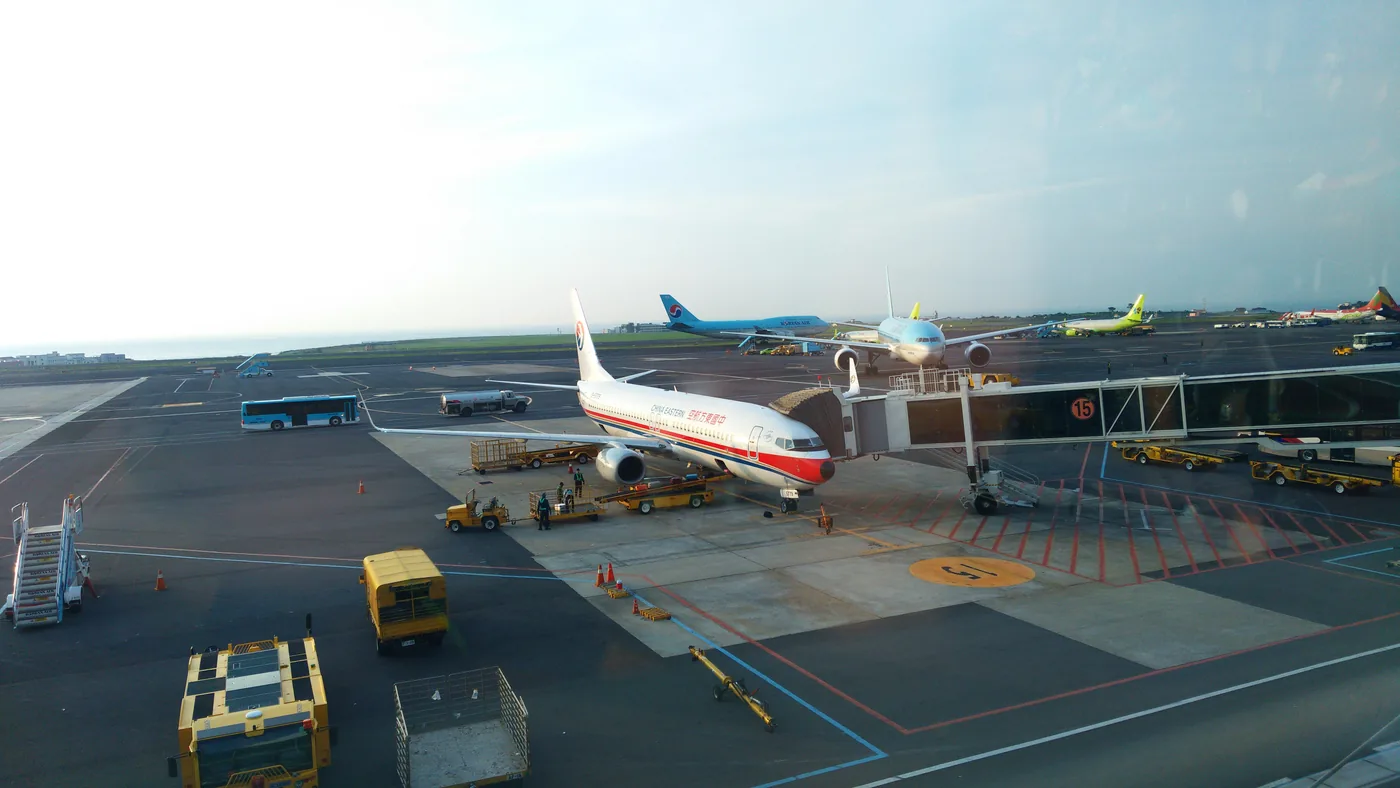 從航廈玻璃窗往下看停機坪,15號空橋旁停著中國東方航空客機,周圍有行李車和車輛,遠處有其他客機與大海 從航廈玻璃窗往下看停機坪,15號空橋旁停著中國東方航空客機,周圍有行李車和車輛,遠處有其他客機與大海