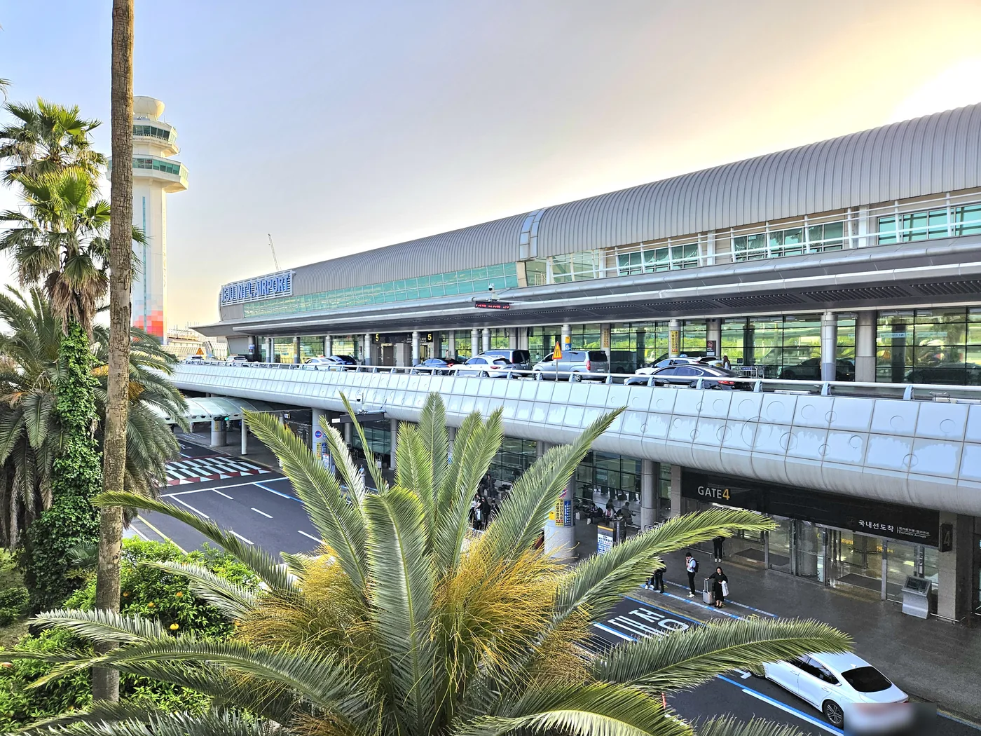 黃昏時斜看濟州國際機場航廈正面,銀色弧形屋頂、左上方的JEJU INT'L AIRPORT招牌、上層高架出發車道上的轎車、下層入口的GATE 4與國內線抵達標示,前景是大片棕櫚葉 黃昏時斜看濟州國際機場航廈正面,銀色弧形屋頂、左上方的JEJU INT'L AIRPORT招牌、上層高架出發車道上的轎車、下層入口的GATE 4與國內線抵達標示,前景是大片棕櫚葉