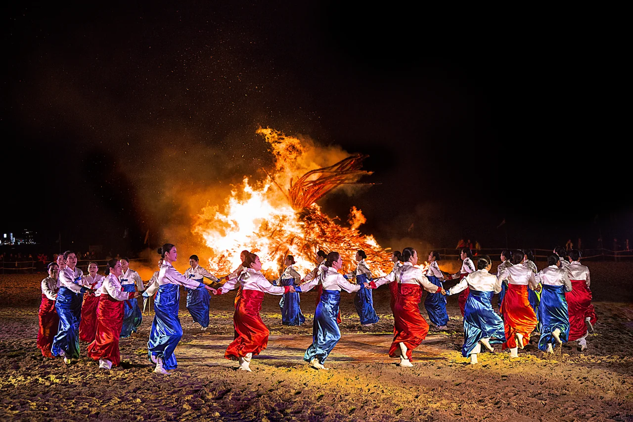 At night on a sandy beach, women in hanbok hold hands and dance in a circle in front of a burning moon house