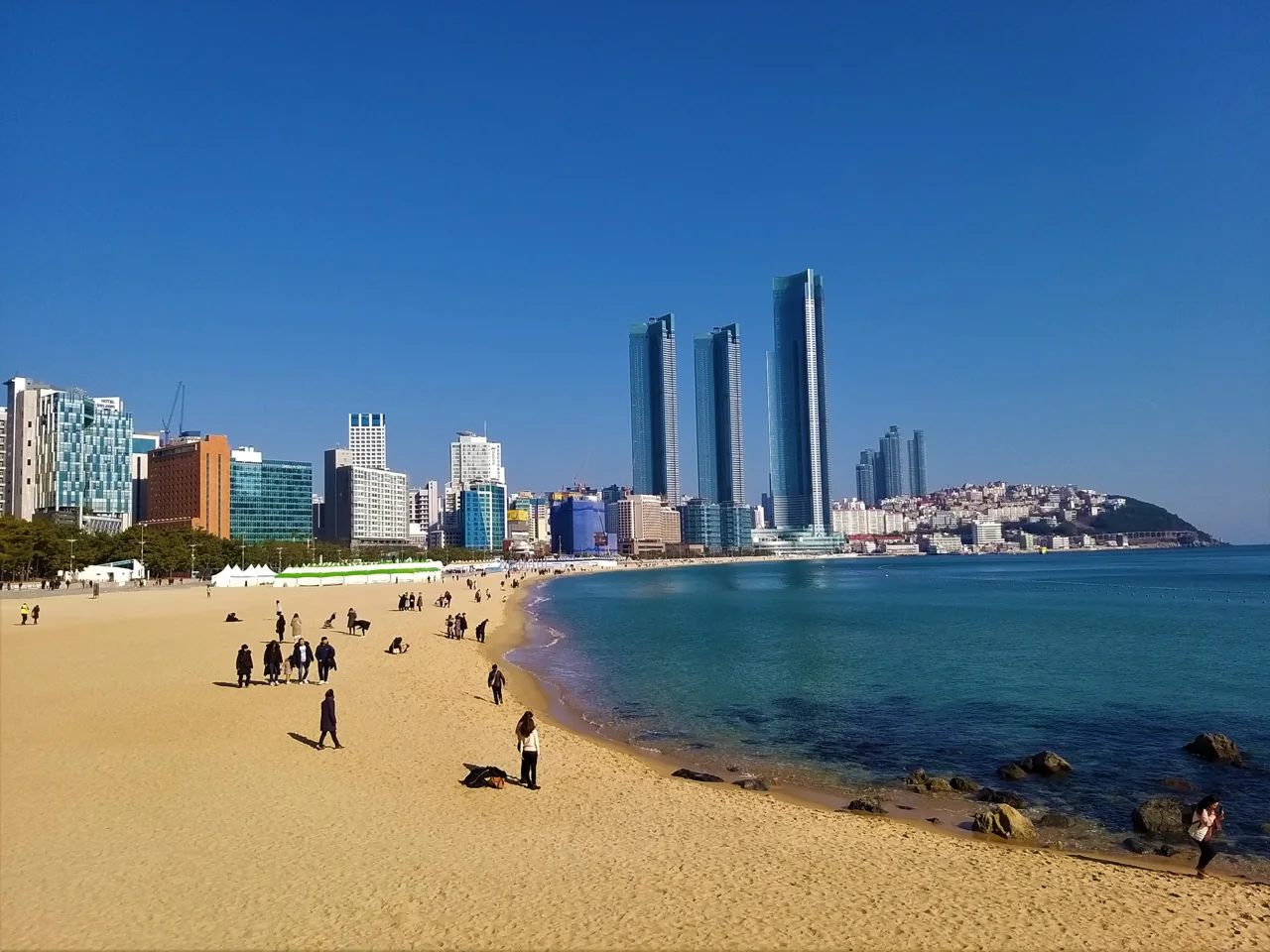 Haeundae Beach seen from the Dongbaekseom side, with people walking on the sand under a clear blue sky and high-rise towers behind