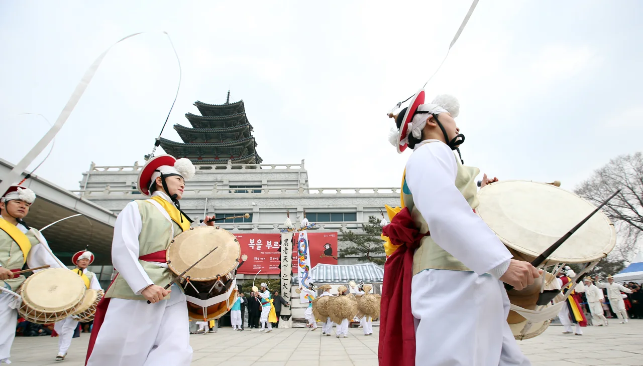 Pungmul musicians in white clothes and sangmo hats beating buk drums as they circle a courtyard, with a pagoda-shaped building behind
