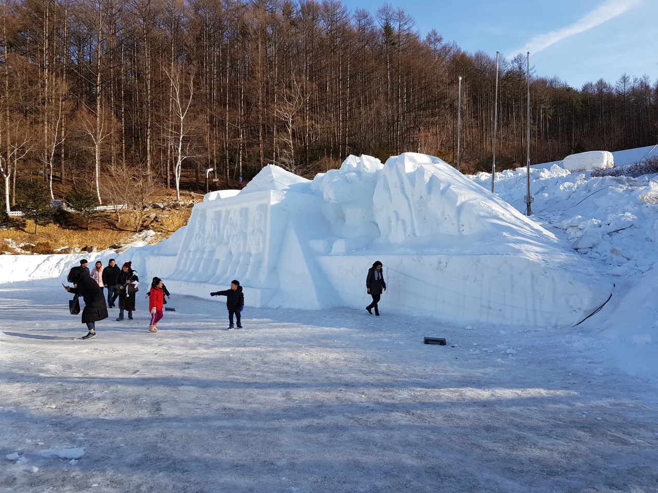 Visitors taking photos in front of a long, tall wall of snow sculptures, with trees and a blue sky behind