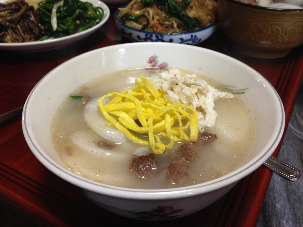 A bowl of tteokguk with diagonally sliced rice cakes, strips of yellow egg and a meat topping